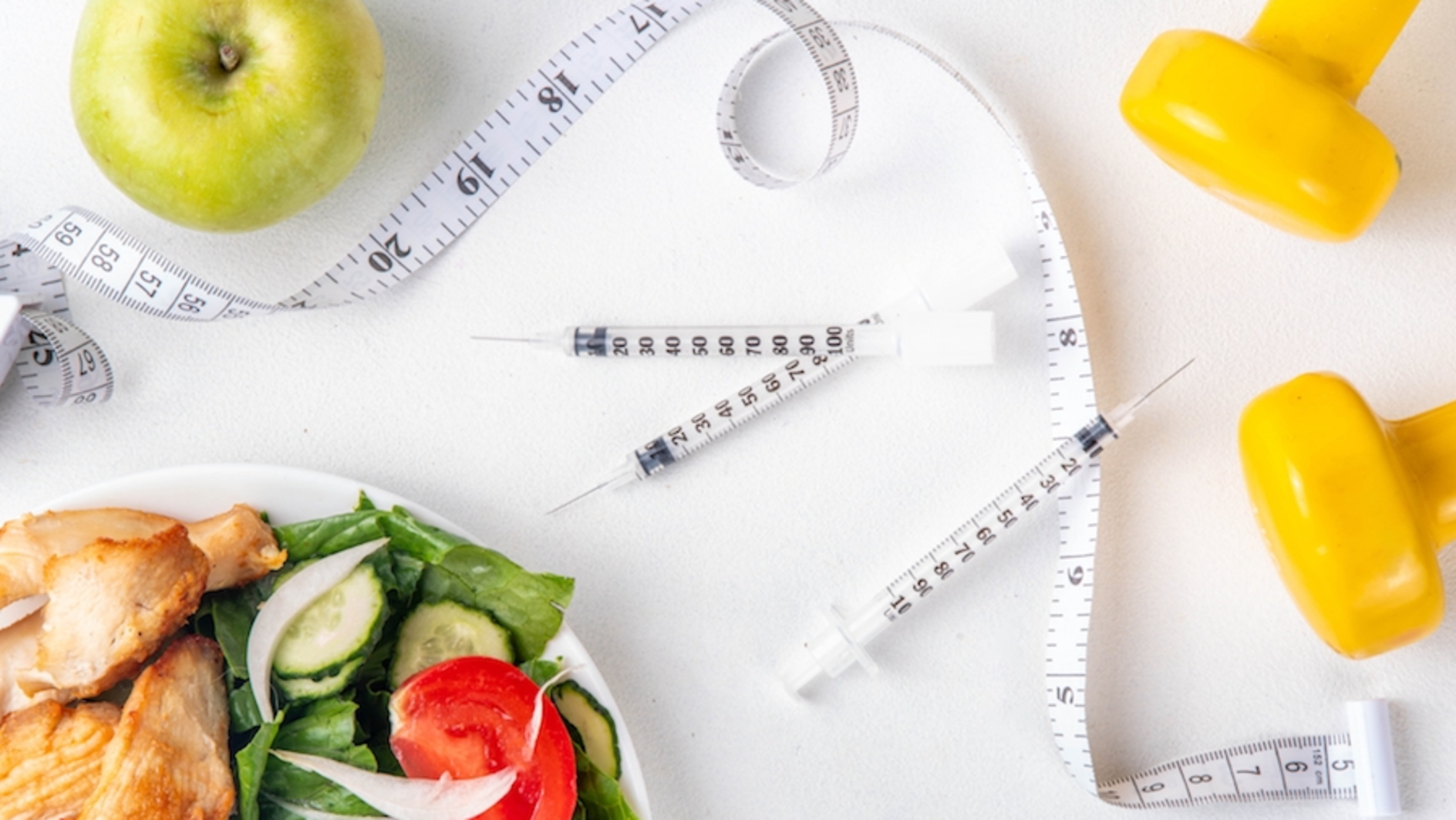 A plate of chicken and greens, an apple, yellow dumbbells, syringes, and a measuring tape sitting on a white background