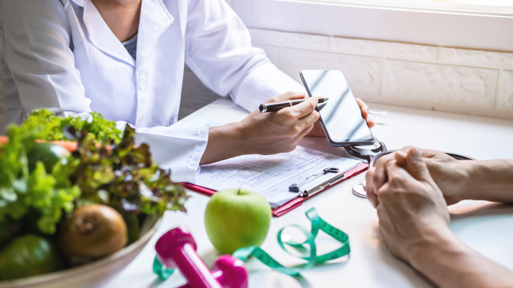 A person in a lab coat explaining something on a smartphone to another person across a desk, with paperwork sitting on the table between them and a bowl of produce and small pink dumbells on the table nearby