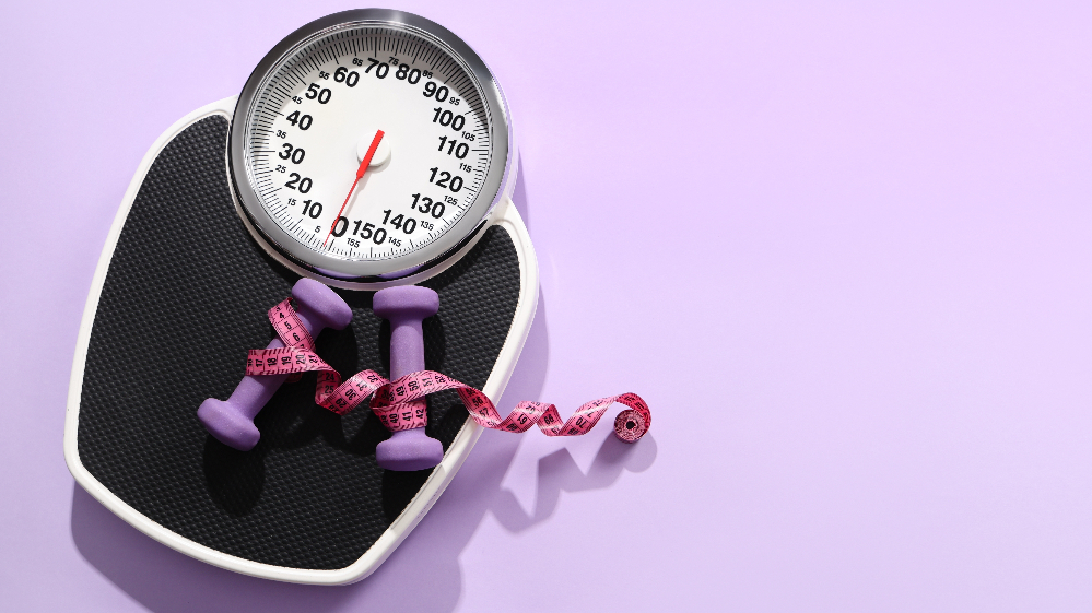 A scale with dumbbells and a measuring tape resting on it, on a lilac background