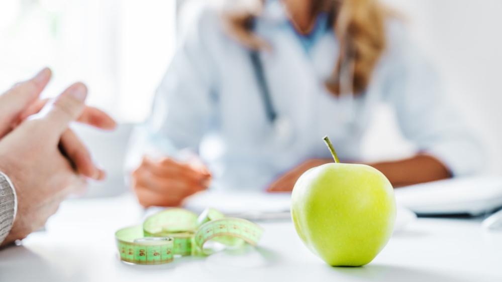 A client talking with a weight loss professional, with an apple and measuring tape on the desk