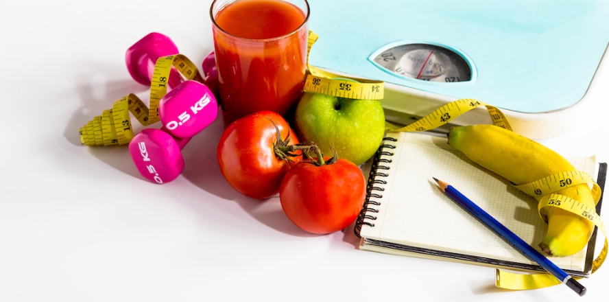 A selection of fruits and vegetables arranged around weights, a scale, and a notebook on a white background