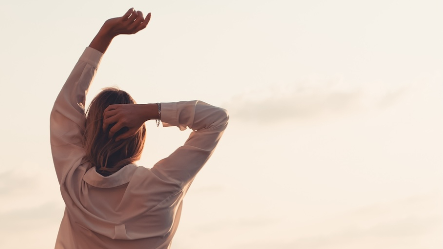 A woman in a pale shirt stretching her arms above her head at sunrise